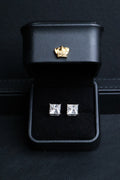 A pair of square-cut stud earrings with clear gemstones in silver settings, presented in an open black jewelry box with a gold crown emblem on the inside lid.