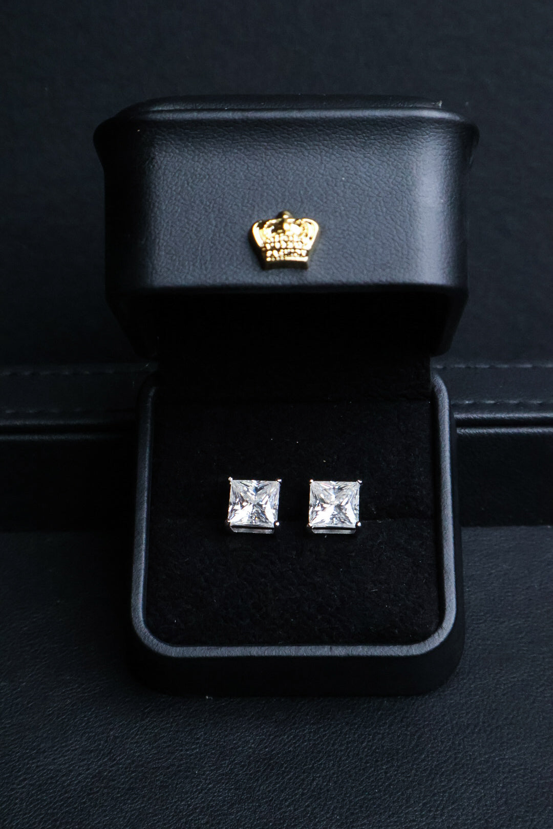 A pair of square-cut stud earrings with clear gemstones in silver settings, presented in an open black jewelry box with a gold crown emblem on the inside lid.